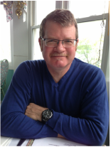 Portrait of a man with glasses in a blue sweater sitting at a table by a sunny window, looking at the camera and smiling lightly.