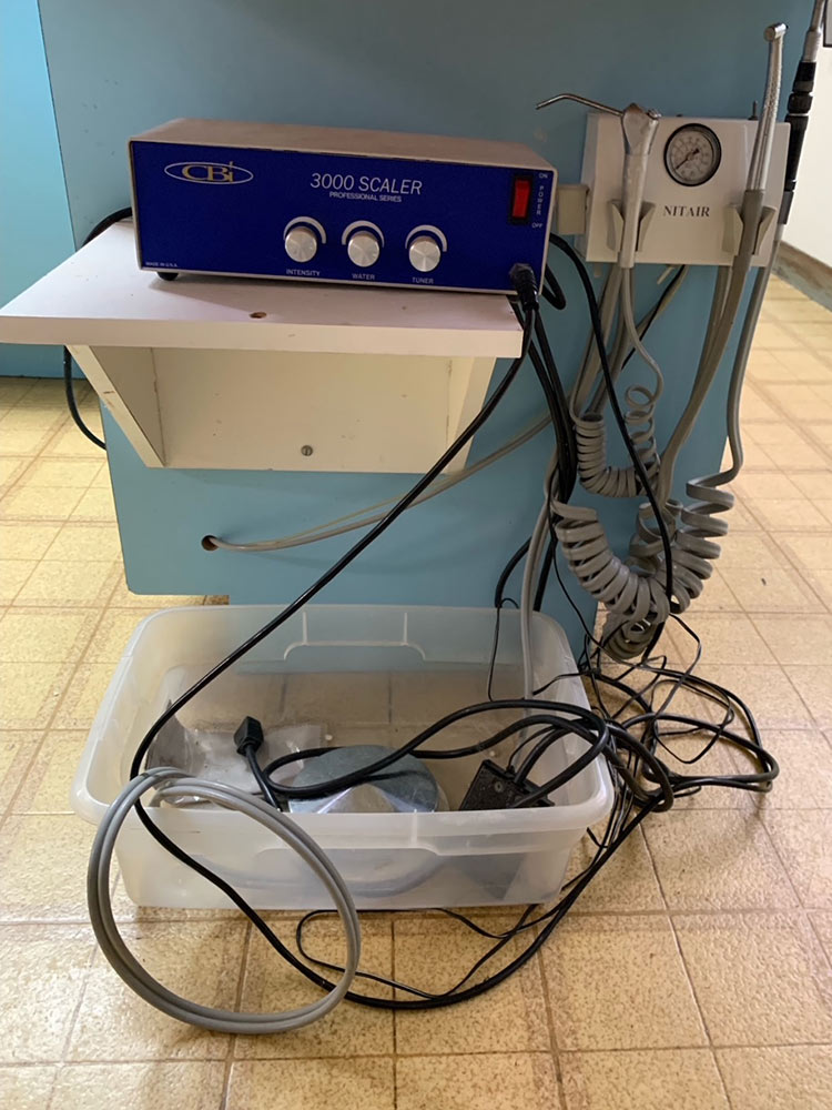 Blue 3000 Scaler dental unit on a shelf with three knobs (Intensity, Water, Tuner) connected to a wall-mounted control; tangled cords and a clear bin with dental tools sit on the floor nearby.
